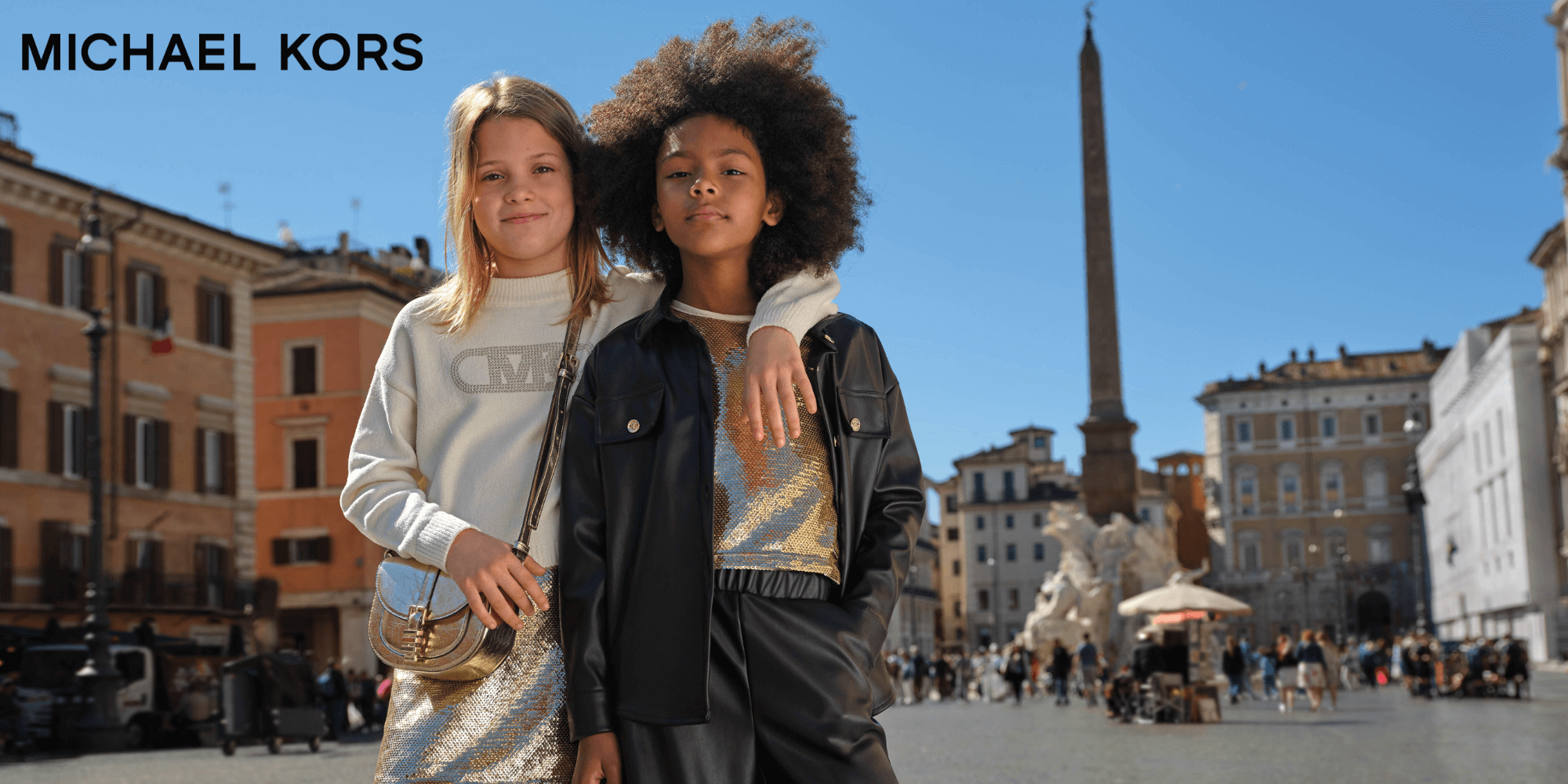 Two young women posing in a historic square with 'Michael Kors' branding.
